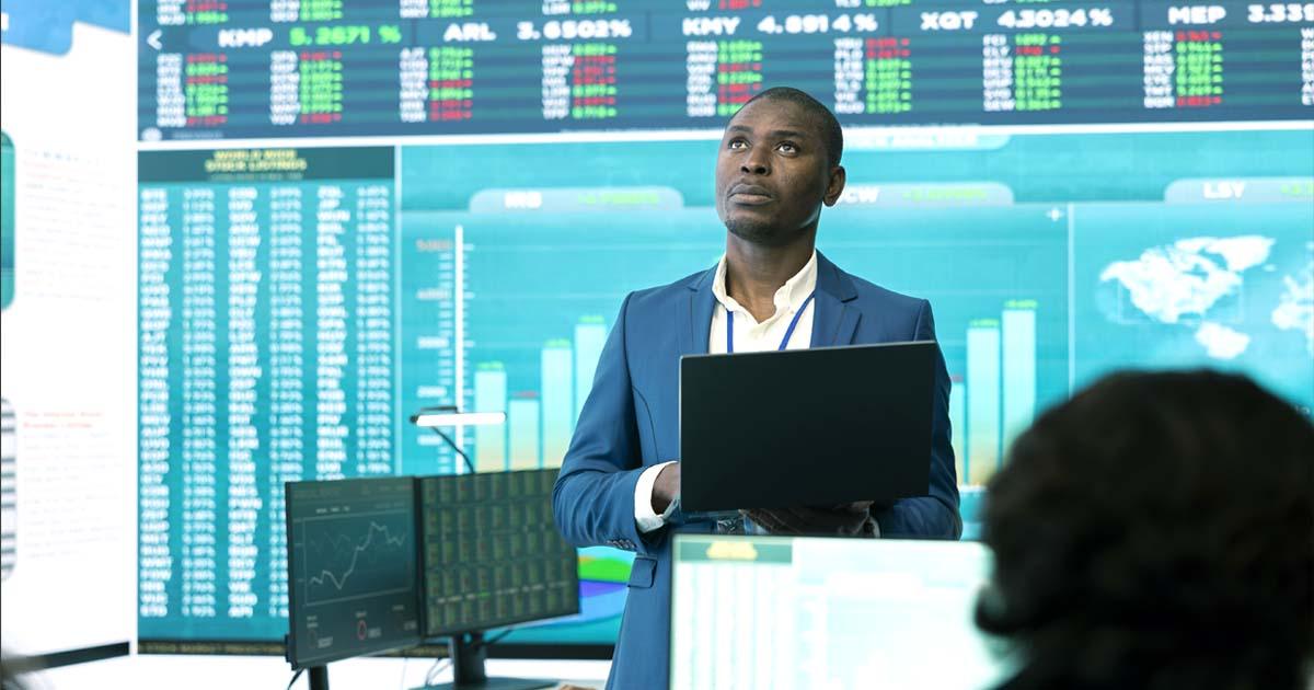 Day Trading: Trader em um ambiente corporativo analisando gráficos financeiros em um laptop, com telas exibindo cotações e dados do mercado.
